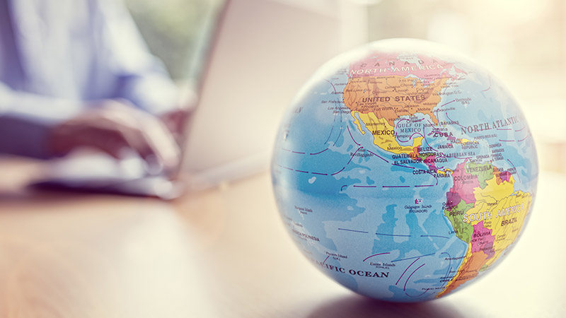 A globe in the foreground, with a computer and hands in the background, out of focus