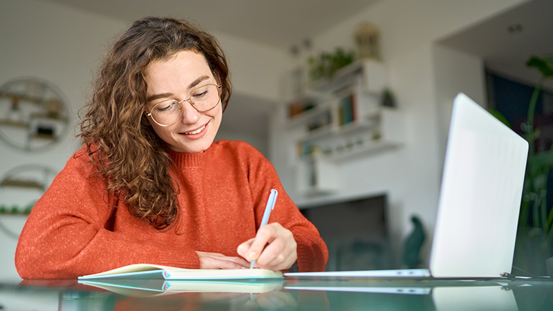 A young woman writing on a piece of paper, with a computer on the desk