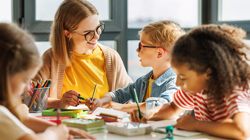 Maestra trabajando con un grupo de niños en una mesa, que contiene lapices de colores y libretas